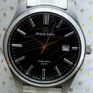 Peut inclure: Une montre Grand Seiko en acier inoxydable avec un cadran noir et des aiguilles argentées. Le cadran affiche la date et le texte "SPRING DRIVE 5 DAYS". La montre a un bracelet argenté et est posée sur un fond blanc.