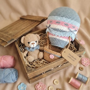 Op de afbeelding: Een handgemaakte haakset met een teddybeer en een heteluchtballon in een houten kist. De ballon is blauw, roze en wit. De teddybeer heeft een blauwe kraag. De kist heeft een label met "Handmade with Love".