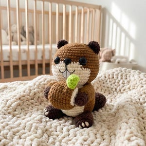 May include: A crocheted beaver toy in brown, cream, and dark brown. The beaver has large black eyes and holds a small green item. The soft toy is placed on a cream-coloured, textured blanket. The toy is handmade.