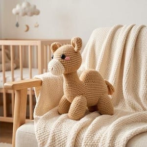 Könnte beinhalten: Ein gehäkeltes Kamelspielzeug in Beige- und Cremetönen sitzt auf einer cremefarbenen Decke auf einem Holzstuhl. Das Kamel hat schwarze Augen, rosa Wangen und eine cremefarbene Schnauze. Ein Kinderbett und ein Wolkenmobile sind im Hintergrund zu sehen.