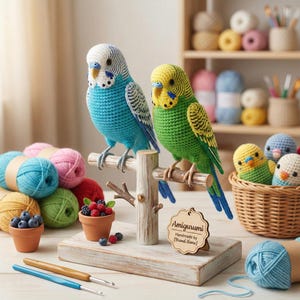 May include: Two crocheted parakeets, one blue and one green, sit on a wooden perch. The blue bird has a white head, the green one a yellow head. The display includes small pots of berries and a wooden sign. Yarn and crochet hooks are also visible.