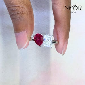 May include: A silver ring with a pear-cut red gemstone and a rectangular, clear gemstone. The ring is held between fingers, highlighting the contrasting colours and shapes of the stones. The brand name "NEOR LUXE" is visible in the upper right corner.