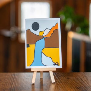 May include: Abstract landscape art print on a small wooden easel. The artwork features a black sun, brown mountains, a blue river, and yellow and orange accents. The art print is displayed on a wooden table.