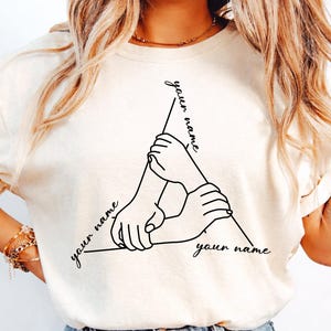 May include: Cream-colored t-shirt featuring a black line art design of three hands clasped together, forming a triangle. The words "your name" are written along each side of the triangle in a script font.