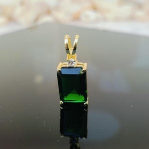 Pode incluir: Um pingente de ouro com uma pedra preciosa retangular verde esmeralda. Uma pequena pedra preciosa transparente é colocada acima da pedra verde. O pingente tem uma argola de ouro para prender a uma corrente.