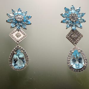 Puede incluir: Un par de pendientes elegantes con un diseño floral y gemas azul claro. Cada pendiente tiene una parte superior en forma de flor, una sección media en forma de diamante y una parte inferior en forma de lágrima, todo en metal plateado.