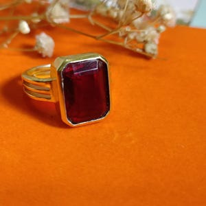 Pode incluir: Um anel dourado com uma grande pedra preciosa retangular vermelha escura. O anel tem uma banda simples com três linhas paralelas e uma moldura retangular. O fundo é laranja vibrante.