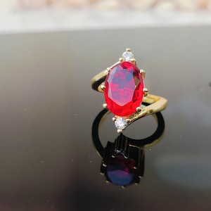 May include: A gold ring with a large, oval-cut, bright red gemstone. The ring is embellished with smaller, clear gemstones on either side of the main stone, adding sparkle. The ring's band has a distinctive, flowing design, creating an elegant look.
