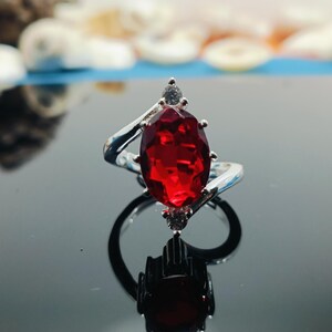 Peut inclure: Bague en argent ornée d'une grosse pierre rouge vif de forme ovale. Le corps de la bague présente un motif tourbillonnant, et de petites pierres d'accent sont serties près de la pierre principale. La bague est présentée sur une surface réfléchissante.