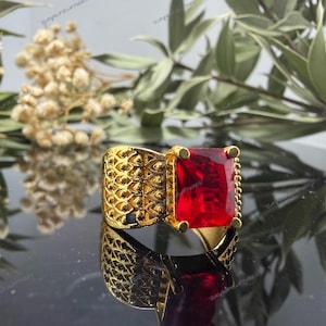 May include: A gold-colored ring with a large, rectangular, red gemstone. The ring has an ornate, textured band with a repeating pattern. The gemstone is set in a square frame, and the ring is displayed against a dark background.
