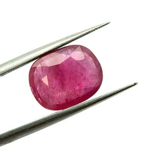 Puede incluir: Una piedra preciosa facetada de forma ovalada, de un vibrante color rojo rosado, sostenida por unas pinzas de metal plateado. La piedra muestra una calidad translúcida con inclusiones internas visibles y una superficie pulida.