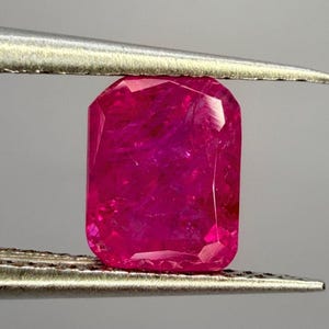 May include: A vibrant, rectangular-cut gemstone with a deep magenta-pink hue and faceted surfaces, held securely between the two metal tips of a pair of tweezers. The stone displays internal clarity and light reflection across its polished faces.