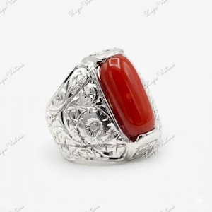 May include: A silver ring with an oval, bright red stone. The ring's band features intricate floral and scrollwork designs. The setting is a bright, polished silver.