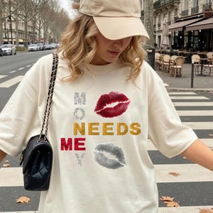 Puede incluir: Una camiseta de manga corta color crema con el texto "MONEY NEEDS ME" dispuesto vertical y horizontalmente, junto con un estampado de labios rojos brillantes y un estampado de labios plateados brillantes. Gorra de béisbol beige y bolso negro.