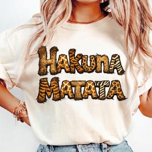 Peut inclure: T-shirt crème avec l'inscription "Hakuna Matata" en marron, avec un motif animalier. Les lettres ont une texture poilue et un mélange de motifs girafe, zèbre et tigre. Le t-shirt est de style décontracté.