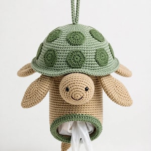 May include: A crochet turtle-shaped plastic bag dispenser in beige and sage green. The turtle has a rounded shell with dark green circular patches, a smiling face with black bead eyes, and a hanging loop at the top for wall mounting.