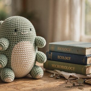 May include: A sage green and cream crocheted plush toy with a rounded body, multiple small limbs, and embroidered facial features, positioned next to a stack of three books titled Biology, Ecology, and Microbiology, and a small potted succulent.