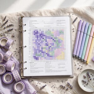 May include: A ring-bound planner featuring a grid page with purple, green, pink, and yellow colored squares. The planner is surrounded by several rolls of patterned washi tape, a set of pastel markers, and dried lavender sprigs.