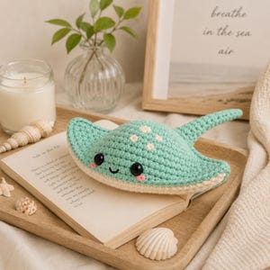 May include: A mint green crocheted stingray plush toy with a long tail, black plastic eyes, small pink embroidered cheeks, and a white underside. The stingray features white embroidered spots on its back and is resting on an open book.