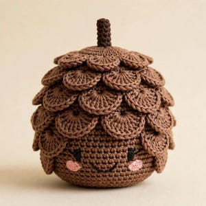 May include: A brown crocheted acorn toy featuring a textured cap made of layered circular stitches. The base is smooth with an embroidered face consisting of two black eyes, a small curved mouth, and two light pink circular cheeks.