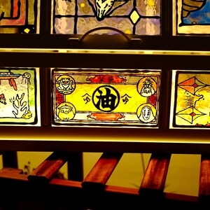 May include: A wooden framed stained glass panel featuring colorful illustrations. The artwork depicts characters and scenes from an animated film. The glass is illuminated from behind, creating a warm, inviting glow. The frame is dark brown.