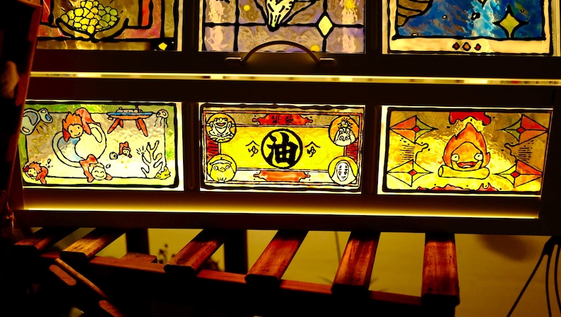 May include: A wooden framed stained glass panel featuring colorful illustrations. The artwork depicts characters and scenes from an animated film. The glass is illuminated from behind, creating a warm, inviting glow. The frame is dark brown.