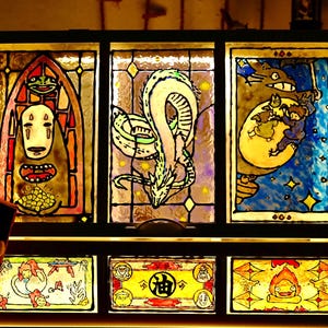 May include: A stained glass panel featuring characters from animated films. The top row depicts a character with a white mask, a dragon, and Totoro. The bottom row shows additional characters. The artwork is framed in black.