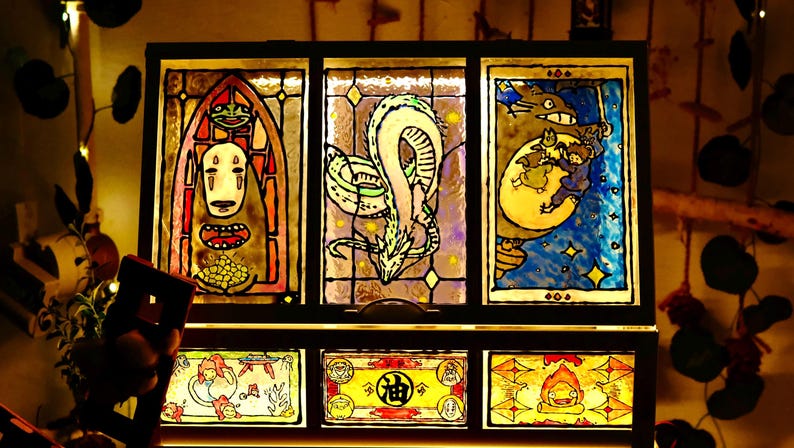 May include: A stained glass panel featuring characters from animated films. The top row depicts a character with a white mask, a dragon, and Totoro. The bottom row shows additional characters. The artwork is framed in black.