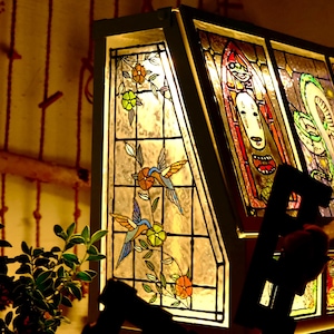 May include: A stained glass lamp with a dark frame. The lamp features colorful panels with images of birds, flowers, and abstract figures. The lamp is illuminated, casting a warm glow. The lamp is a decorative home decor item.