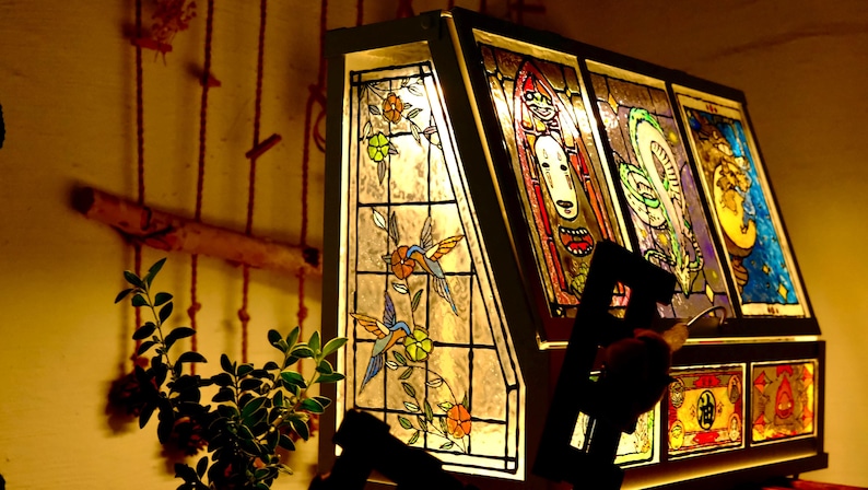 May include: A stained glass lamp with a dark frame. The lamp features colorful panels with images of birds, flowers, and abstract figures. The lamp is illuminated, casting a warm glow. The lamp is a decorative home decor item.