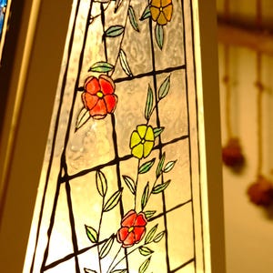 May include: Close-up of a stained glass lamp with a floral design. The glass features red, yellow, and orange flowers with green leaves, outlined in black. The lamp has a white frame and is illuminated from within.