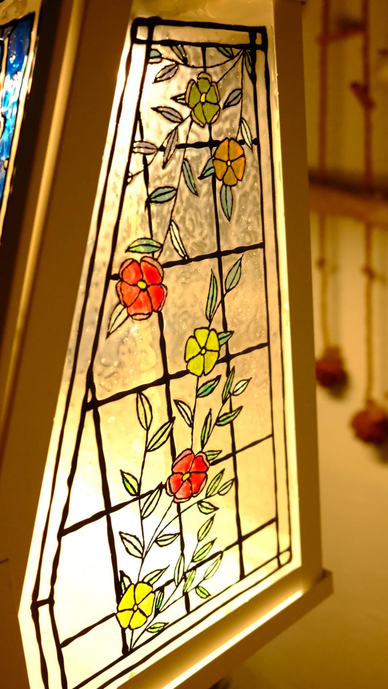 May include: Close-up of a stained glass lamp with a floral design. The glass features red, yellow, and orange flowers with green leaves, outlined in black. The lamp has a white frame and is illuminated from within.