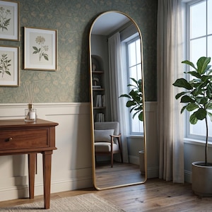 May include: A tall, arched-top mirror with a gold-colored frame. The mirror reflects a room with a window, a chair, and a potted plant. The mirror is standing on a wooden floor.