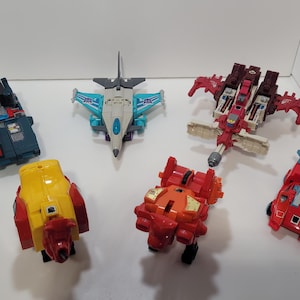 May include: A collection of colorful toy robots in various forms, including a teal and red truck, a yellow and red car, a red and white jet, and a red and black robot. The toys are arranged on a white surface.