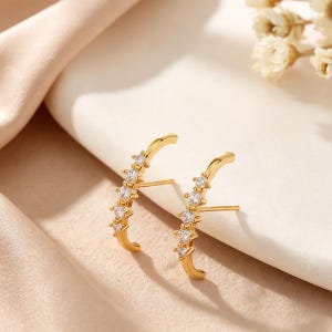 May include: A pair of gold-toned ear climbers featuring a curved bar design. Each earring is set with five clear, round-cut stones arranged in a row along the metal arc, with a straight post extending from the back for secure attachment to the ear.