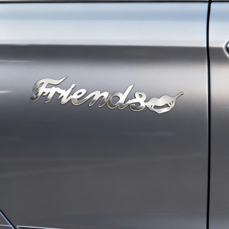 May include: A chrome-colored "Friends" emblem, affixed to a gray vehicle door. The word "Friends" is written in a cursive font, with a stylized design of a mouth and lips at the end of the word.