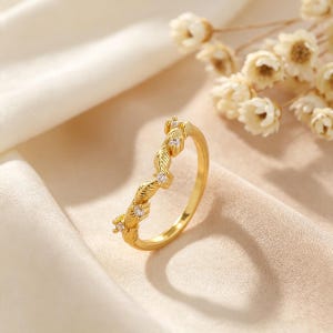 Op de afbeelding: Een goudkleurige ring in chevronvorm met een reeks getextureerde, bladachtige segmenten. Kleine, heldere, rond geslepen stenen zijn in de metalen structuur gezet, afwisselend tussen de bladmotieven langs de bovenste helft.