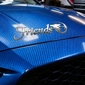 May include: A close-up of a blue carbon fibre car bonnet with the word "Friends" in silver script. A silver emblem of a stylized dragon is next to the word. The bonnet has a glossy finish.