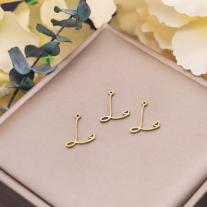 Può includere: Tre ciondoli in metallo color oro a forma di lettera "L". Ogni ciondolo ha un piccolo anello nella parte superiore per il fissaggio ai gioielli. I ciondoli sono esposti su una superficie marrone chiaro.