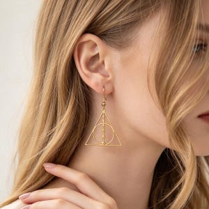 May include: A gold-toned dangle earring featuring a geometric design composed of a triangle enclosing a circle with a vertical line of small spheres running through the centre, suspended from a simple hook wire finding for pierced ears.