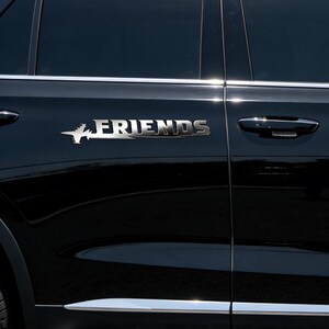May include: A silver car emblem displaying the word "FRIENDS" in a unique font. The emblem is affixed to a black car door. A stylized airplane design precedes the word "FRIENDS".