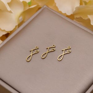 Può includere: Tre ciondoli dorati a forma di lettera "J" con un dettaglio a cuore. Ogni ciondolo ha un piccolo anello nella parte superiore per il fissaggio ai gioielli. I ciondoli sono esposti su una superficie dai toni neutri.