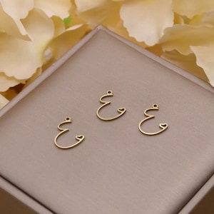 May include: Three gold-colored charms in the shape of the Arabic letter 'ain, each with a small loop at the top for hanging. The charms are displayed on a neutral-toned surface, suggesting jewelry or craft supplies.