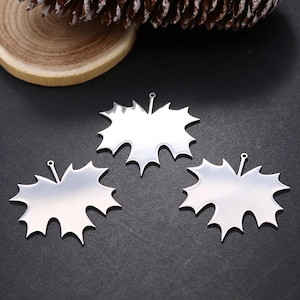 Op de afbeelding: Drie zilverkleurige metalen bedels in de vorm van esdoornbladeren. Elke bedel heeft een glad, reflecterend oppervlak met een kleine ronde lus aan de bovenkant voor bevestiging. Het bladontwerp heeft meerdere puntige lobben en een steeltje.
