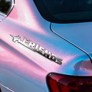 May include: A chrome car emblem displaying the word "FRIENDS" with an airplane design. The emblem is attached to a car with a pearlescent pink and blue paint finish, positioned near the rear light.