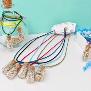 May include: A collection of necklaces featuring small glass vials filled with sand and sealed with cork stoppers. The necklaces have cords in various colors including black, blue, pink, and green, and are displayed with a glass bottle and white boxes.