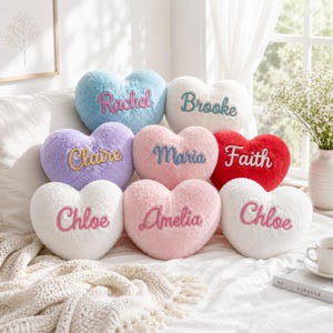Peut inclure: Un ensemble de huit coussins en forme de cœur en peluche de différentes couleurs, notamment bleu, blanc, violet, rose et rouge. Chaque coussin porte un prénom brodé en écriture cursive, soit Rachel, Brooke, Claire, Maria, Faith, Chloe et Amelia.