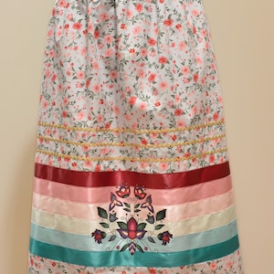 May include: A long skirt with a floral pattern in pink and white. The skirt features horizontal bands of red, pink, and teal ribbons, with a floral design at the bottom. Gold trim accents the design.
