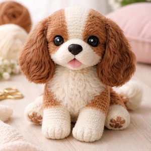 Puede incluir: Un peluche de perro Cavalier King Charles Spaniel tejido a mano. El perro tiene pelaje marrón y blanco, ojos negros grandes y una lengua rosa. El peluche está sentado sobre una superficie clara.