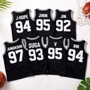 May include: Black basketball jerseys with white trim and lettering. Each jersey displays a name and number: J-HOPE 94, JIMIN 95, JIN 92, JUNGKOOK 97, SUGA 93, V 95, and RM 94. A basketball and net are visible in the background.